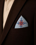 Hand embroidered Kasuti pocket square in 100% Mulmul Khadi featuring a lotus motif rendered in ember red and deep indigo threadwork, styled within the breast pocket of a dark chocolate tailored jacket against a soft ivory shirt. The composition highlights the radial geometry of traditional Kasuti embroidery alongside the fine handwoven texture, hand-finished detailing, and quiet material presence of Mulmul Khadi under warm archival lighting.