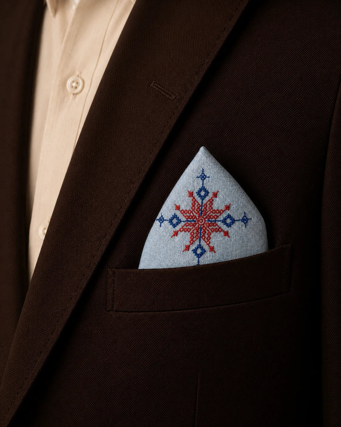 Hand embroidered Kasuti pocket square in 100% Mulmul Khadi featuring a lotus motif rendered in ember red and deep indigo threadwork, styled within the breast pocket of a dark chocolate tailored jacket against a soft ivory shirt. The composition highlights the radial geometry of traditional Kasuti embroidery alongside the fine handwoven texture, hand-finished detailing, and quiet material presence of Mulmul Khadi under warm archival lighting.