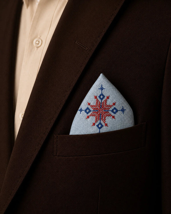 Hand embroidered Kasuti pocket square in 100% Mulmul Khadi featuring a lotus motif rendered in ember red and deep indigo threadwork, styled within the breast pocket of a dark chocolate tailored jacket against a soft ivory shirt. The composition highlights the radial geometry of traditional Kasuti embroidery alongside the fine handwoven texture, hand-finished detailing, and quiet material presence of Mulmul Khadi under warm archival lighting.