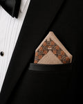Hand embroidered Ritam Kasuti pocket square in 100% Mulmul Khadi featuring a Swastik geometric border composition rendered in burnished copper and deep espresso threadwork, folded within the breast pocket of a black tuxedo jacket paired with a pleated ivory shirt. The composition highlights the formal discipline of counted Kasuti embroidery, softened Mulmul weave texture, and the contrast between ceremonial tailoring and enduring handcraft under softened ambient light.
