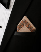 Hand embroidered Ritam Kasuti pocket square in 100% Mulmul Khadi featuring a Swastik geometric border composition rendered in burnished copper and deep espresso threadwork, folded within the breast pocket of a black tuxedo jacket paired with a pleated ivory shirt. The composition highlights the formal discipline of counted Kasuti embroidery, softened Mulmul weave texture, and the contrast between ceremonial tailoring and enduring handcraft under softened ambient light.