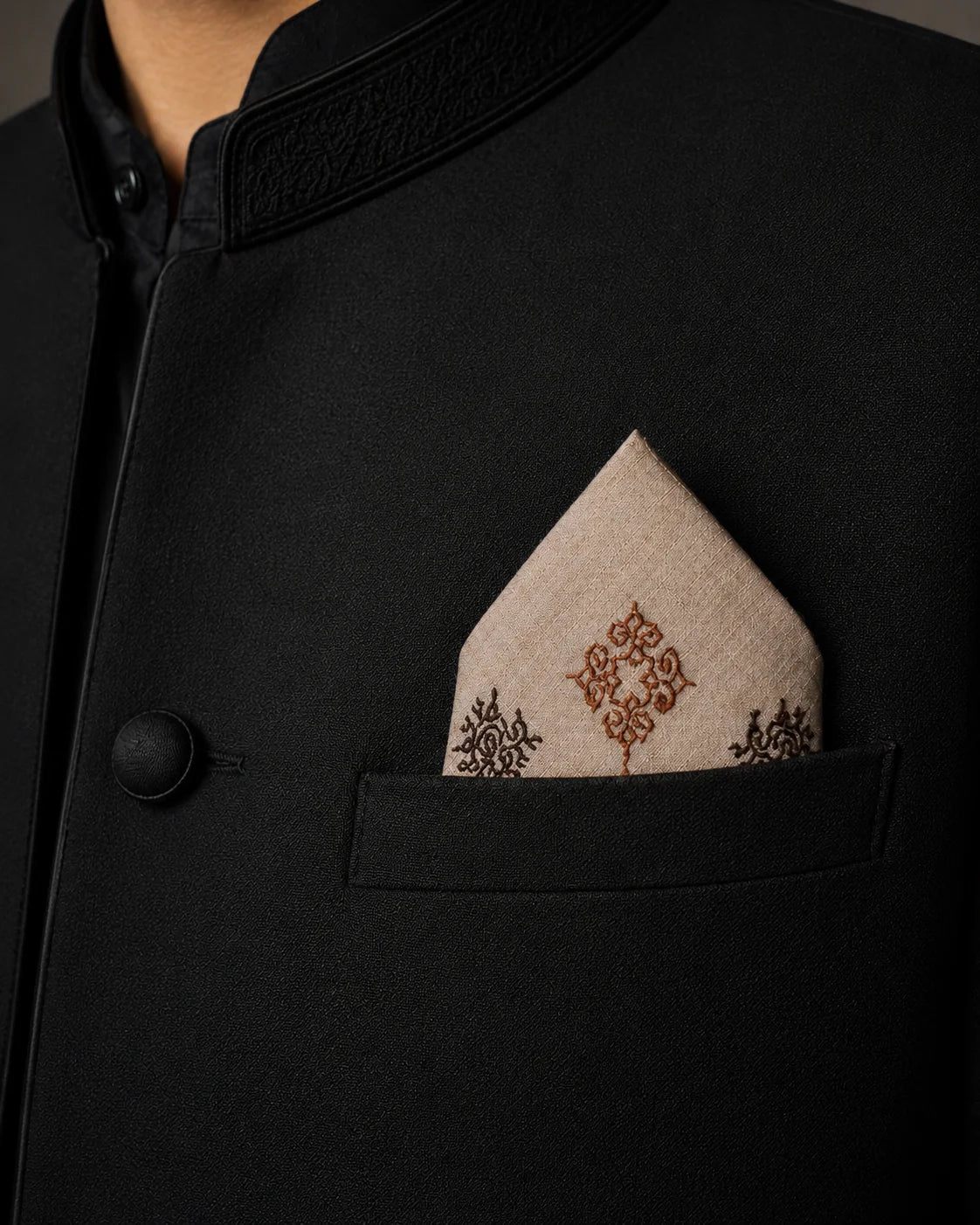 Tarang hand embroidered Kasuti pocket square in 100% Mulmul Khadi featuring peacock and interconnected geometric motifs rendered in burnt copper and deep espresso threadwork. Styled within the breast pocket of a black Nehru jacket, the image emphasizes the quiet precision of counted Kasuti embroidery, the softened texture of handwoven Mulmul Khadi, and the restrained elegance of ceremonial Indian tailoring under low directional lighting.
