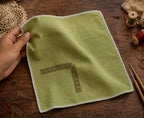 Kasuti hand embroidered pocket square on 100% Mulmul Khadi featuring a Swastik motif executed through traditional Murgi and Gavanti stitch techniques, finished with hand-crocheted edging and photographed on a worn wooden textile surface alongside loose embroidery threads, crochet lace, wooden-handled tools, and visible hand interaction that reflects the measured cadence of slow embroidery practice carried through generations of Karnataka textile work.