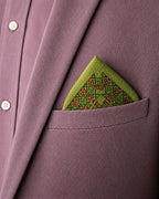 Hand embroidered Vyana Kasuti pocket square in 100% Mulmul Khadi featuring a Swastik motif executed in moss green and deep maroon threadwork, folded within the breast pocket of a muted rose textured jacket layered over a tonal shirt. The arrangement foregrounds the architectural order of counted Kasuti embroidery, the airy translucence of Mulmul Khadi, and the tactile contrast between soft handwoven cloth and structured tailoring under subdued archival lighting.