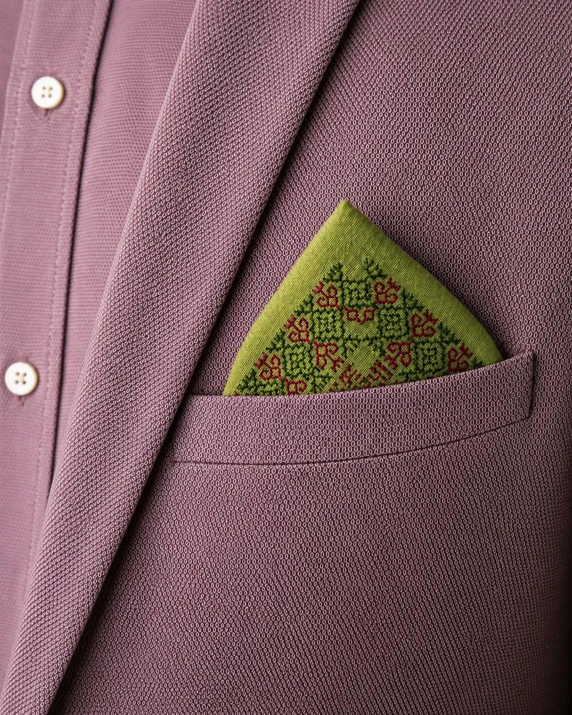 Hand embroidered Vyana Kasuti pocket square in 100% Mulmul Khadi featuring a Swastik motif executed in moss green and deep maroon threadwork, folded within the breast pocket of a muted rose textured jacket layered over a tonal shirt. The arrangement foregrounds the architectural order of counted Kasuti embroidery, the airy translucence of Mulmul Khadi, and the tactile contrast between soft handwoven cloth and structured tailoring under subdued archival lighting.