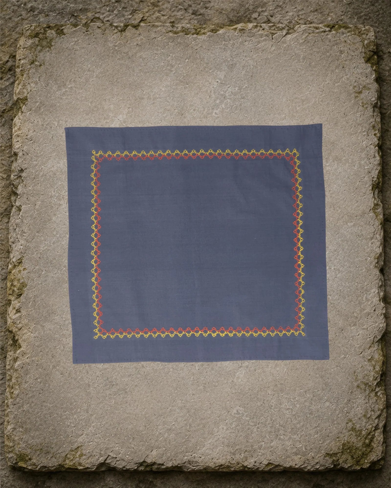 Kasuti Kudremukh hand-embroidered khadi cotton handkerchief with a continuous yellow and orange Betel Box motif border, displayed on a weathered stone surface.