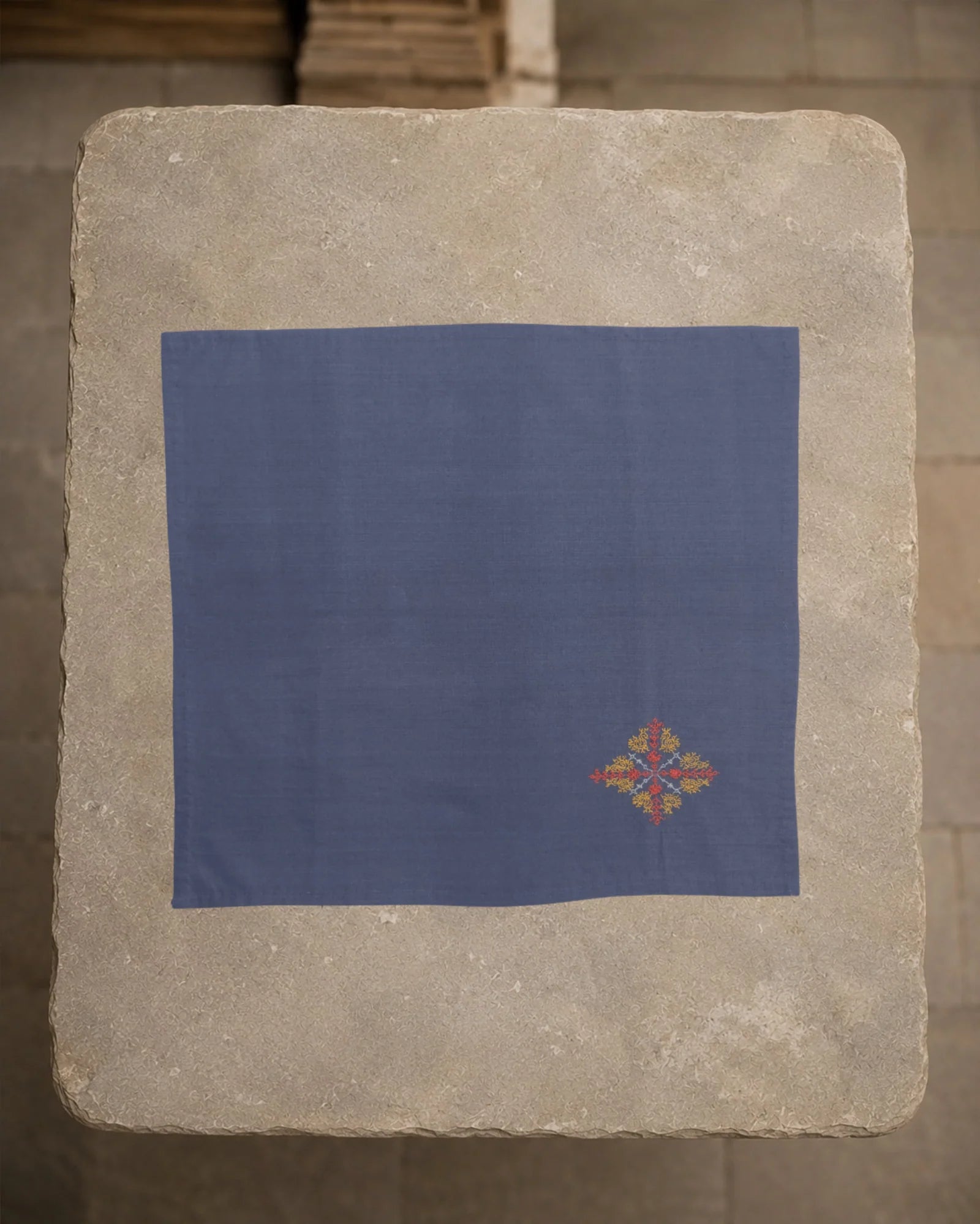 Kasuti Malenadu khadi cotton handkerchief laid open on a weathered stone plinth, with a small hand-embroidered peacock motif positioned near one corner.