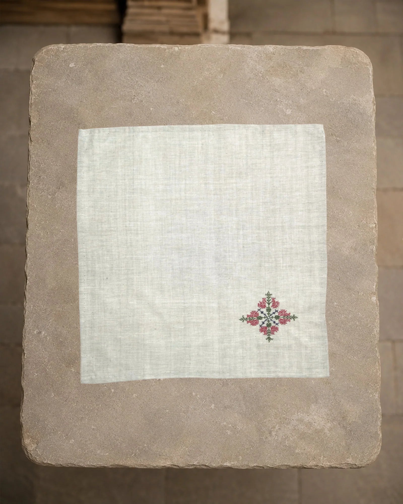Kasuti Mullayanagiri hand-embroidered khadi cotton handkerchief laid open on a weathered stone plinth, with a small peacock motif worked in deep pink, green and blue threads.