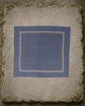 Sharavati Kasuti hand-embroidered khadi cotton handkerchief laid flat on a weathered stone slab, framed by a repeating Betel Box border in white and ochre thread.
