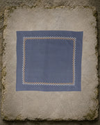 Sharavati Kasuti hand-embroidered khadi cotton handkerchief laid flat on a weathered stone slab, framed by a repeating Betel Box border in white and ochre thread.