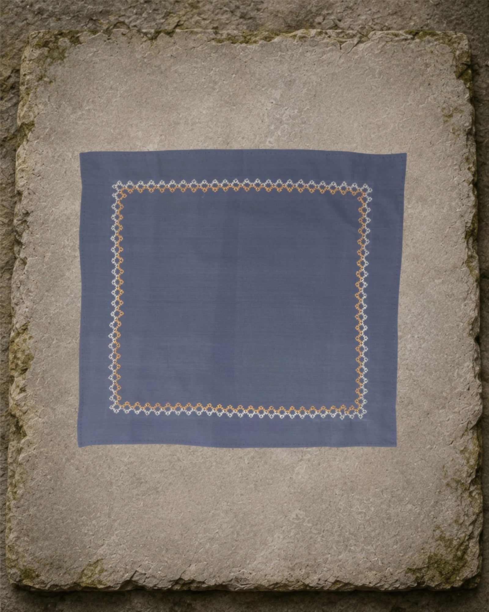 Sharavati Kasuti hand-embroidered khadi cotton handkerchief laid flat on a weathered stone slab, framed by a repeating Betel Box border in white and ochre thread.