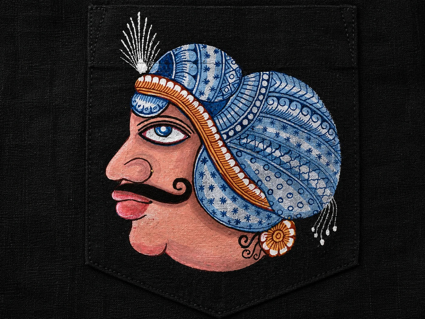 Close-up of hand-painted Odisha Pattachitra king portrait motif on black linen shirt pocket showing fine linework and patterned detailing.