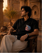 Odisha Pattachitra hand-painted black linen shirt featuring a portrait motif on the chest pocket, observed seated within a sandstone courtyard in warm evening light.