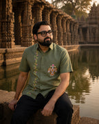 Odisha Pattachitra hand-painted linen shirt featuring intertwined bird forms and lotus elements on the chest pocket and floral vertical detailing along the placket, observed beside a temple pond before carved stone gallery architecture in warm evening light.