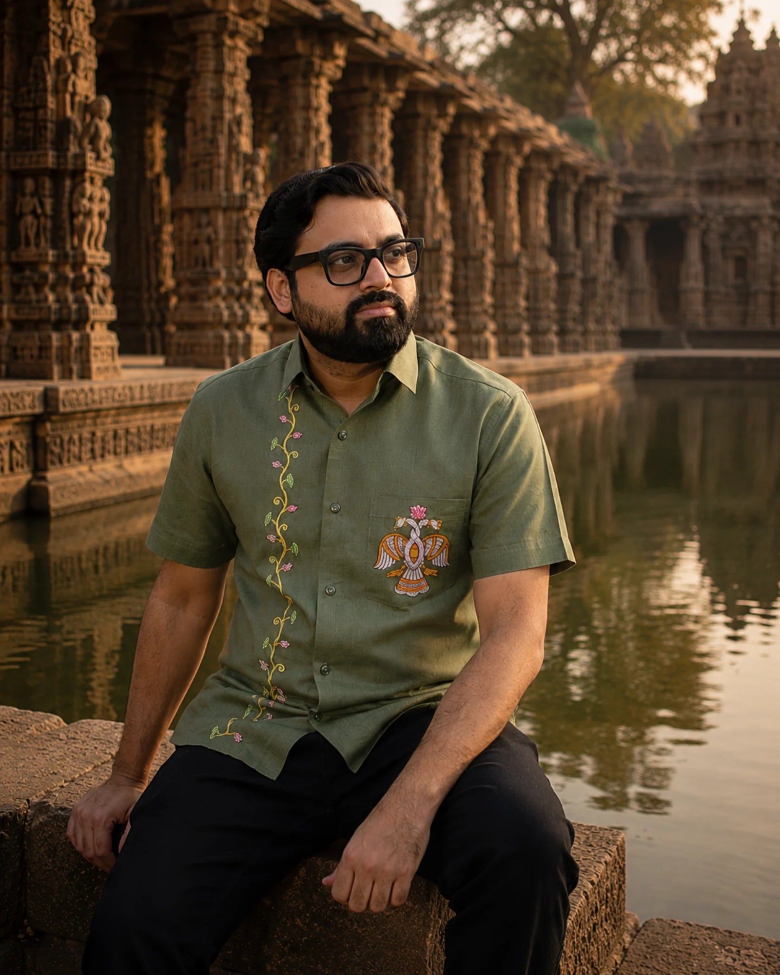 Odisha Pattachitra hand-painted linen shirt featuring intertwined bird forms and lotus elements on the chest pocket and floral vertical detailing along the placket, observed beside a temple pond before carved stone gallery architecture in warm evening light.