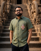 Odisha Pattachitra hand-painted linen shirt featuring intertwined bird forms on the chest pocket and floral vertical detailing along the placket, observed before carved temple architecture in warm evening light.