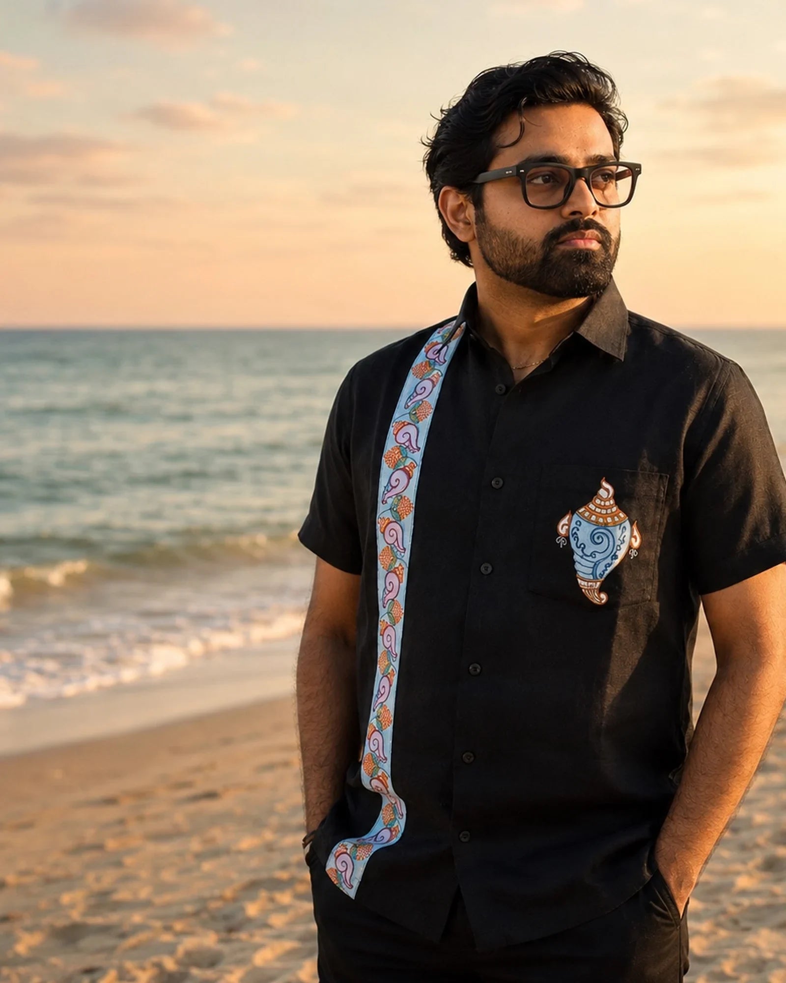 Odisha Pattachitra hand-painted linen shirt featuring a conch artwork on the chest pocket and vertical detailing composed of floral and conch forms along the placket, observed along the seashore in warm sunset light.
