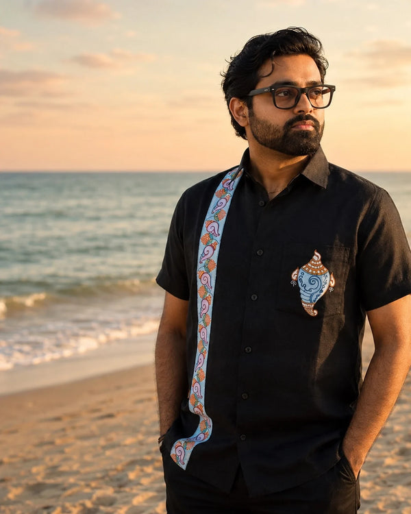 Odisha Pattachitra hand-painted linen shirt featuring a conch artwork on the chest pocket and vertical detailing composed of floral and conch forms along the placket, observed along the seashore in warm sunset light.