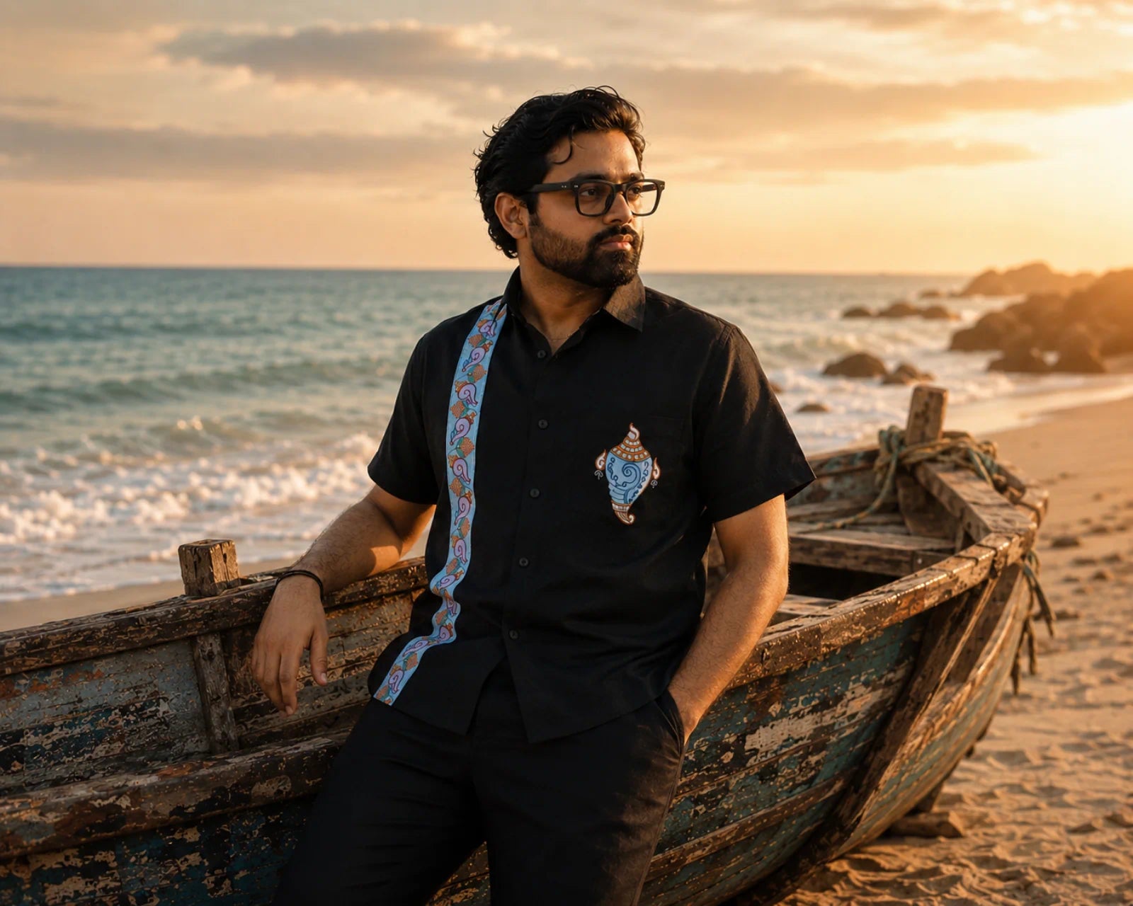 Odisha Pattachitra hand-painted linen shirt featuring a conch artwork on the chest pocket and vertical detailing composed of floral and conch forms along the placket, observed beside a weathered wooden boat along the seashore in warm evening light.