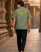 Odisha Pattachitra hand-painted linen shirt featuring twin parrot motifs surrounding a ceremonial kalash artwork across the back, observed walking barefoot through a carved temple gallery in warm archival light with historic stone architecture in the background.