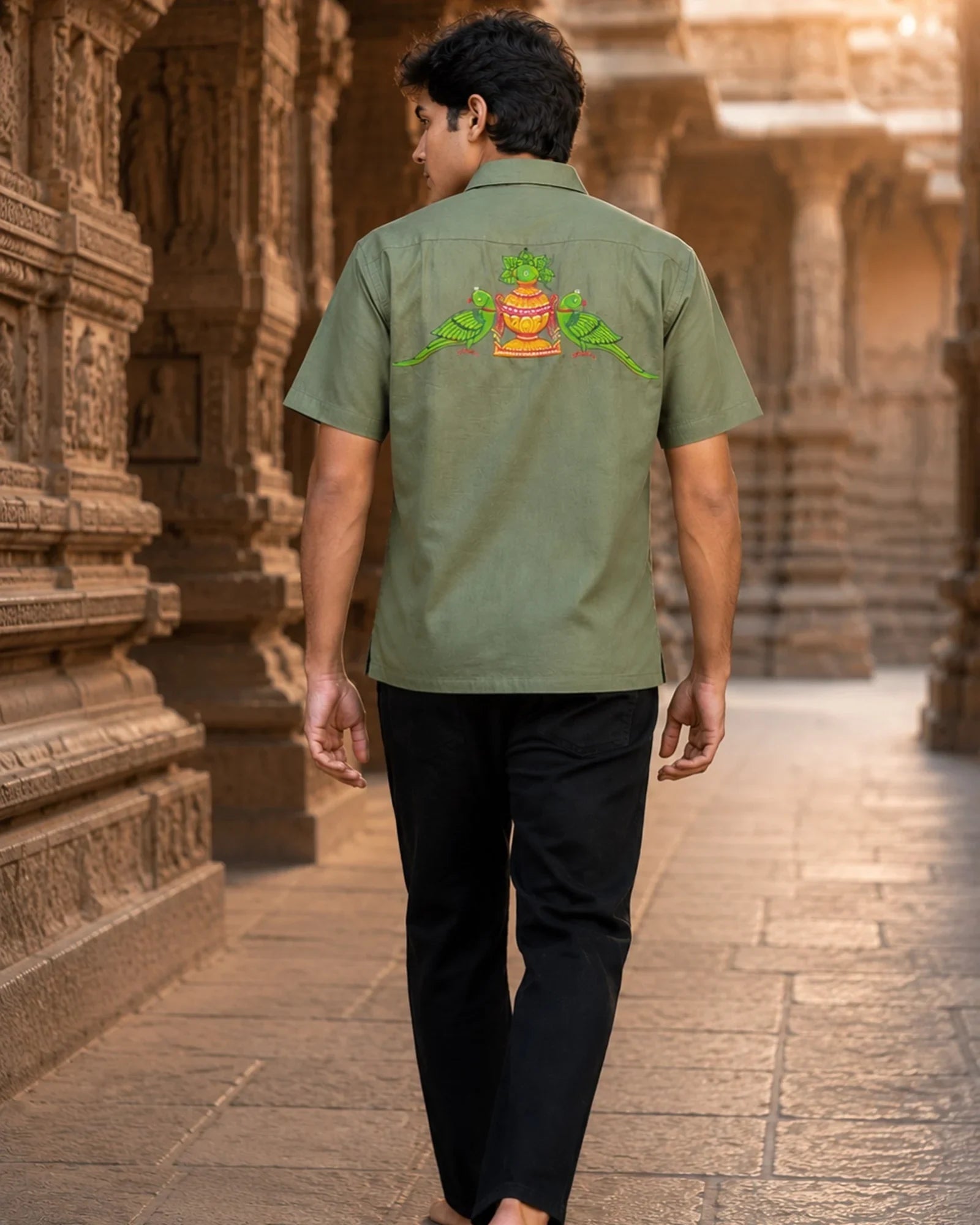 Odisha Pattachitra hand-painted linen shirt featuring twin parrot motifs surrounding a ceremonial kalash artwork across the back, observed walking barefoot through a carved temple gallery in warm archival light with historic stone architecture in the background.