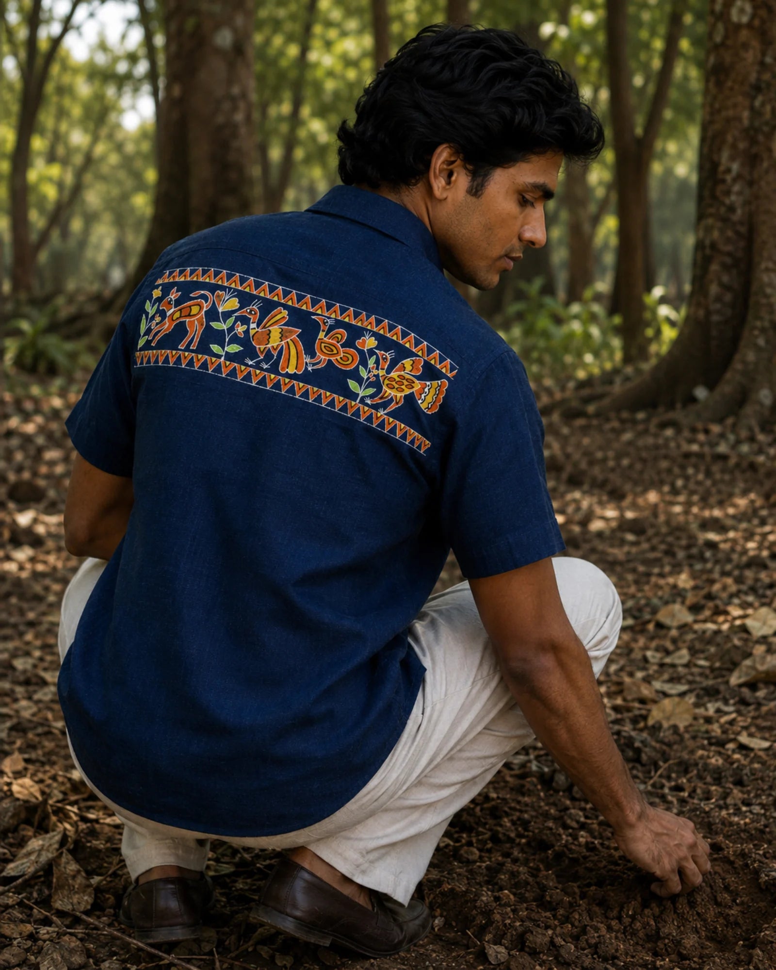 Sohrai Jeev Dhara hand-painted linen shirt viewed from the back in a forest setting, featuring a peacock and peahen, fawn and flowering vegetation within hand-painted borders.