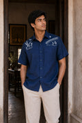 Warli Aavartan hand-painted linen shirt featuring detailed Warli village and Tarpa dance scenes across the shoulders, photographed in an architectural doorway.