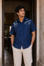 Warli Aavartan hand-painted linen shirt featuring detailed Warli village and Tarpa dance scenes across the shoulders, photographed in an architectural doorway.