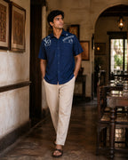 Warli Aavartan hand-painted linen shirt featuring detailed Warli village and Tarpa dance scenes across the shoulders, photographed in a heritage restaurant gallery.