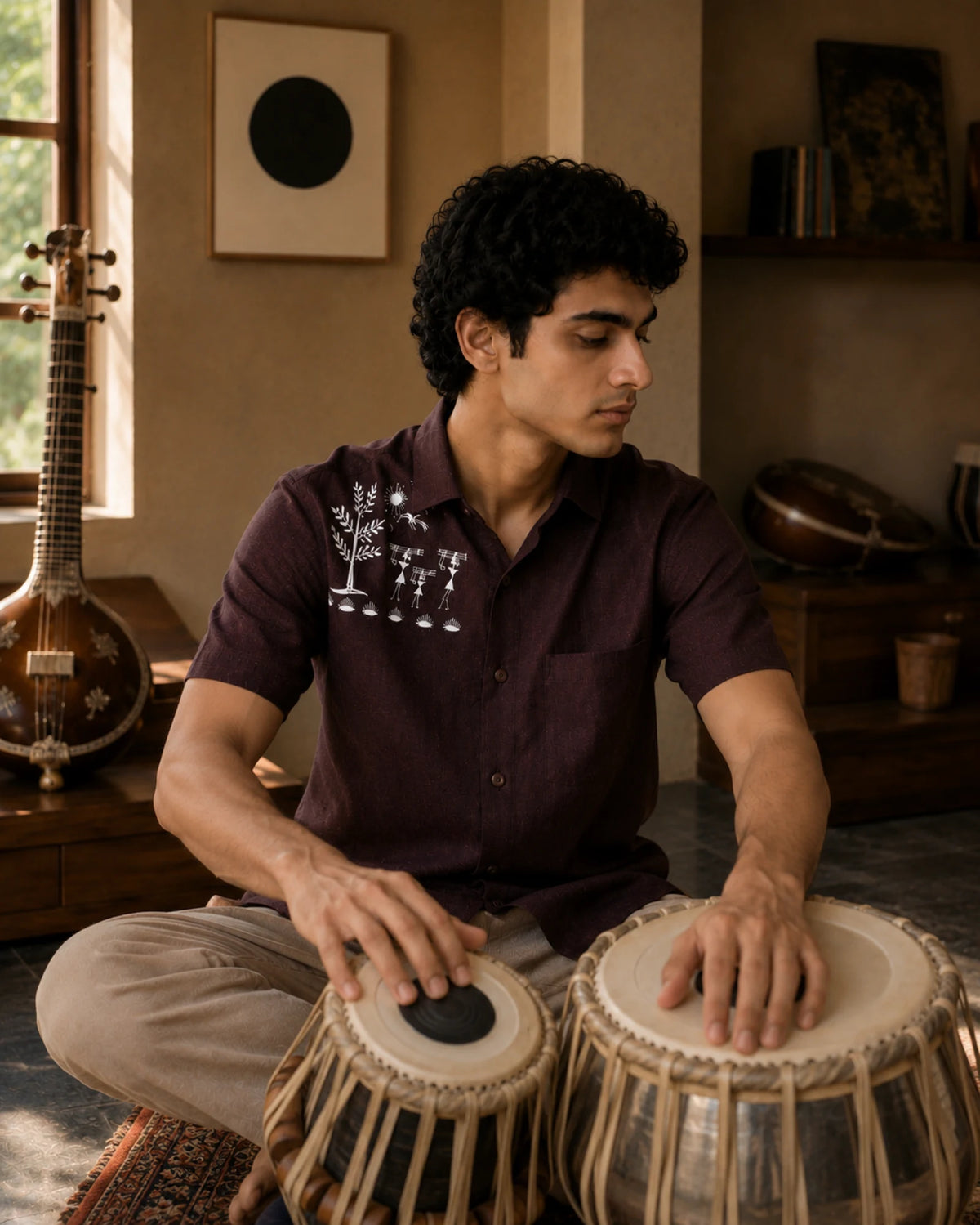 Warli Adiva hand-painted linen shirt styled context featuring shoulder artwork of a tree, birds, sun and figures carrying logs of wood, worn while playing the tabla in a traditional music room.