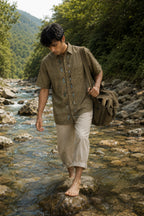 Warli hand-painted linen shirt featuring a vertical narrative of tribal figures, birds and floral motifs along the button placket, observed crossing a shallow forest stream barefoot with a canvas satchel, surrounded by flowing water, river stones and a remote valley landscape.