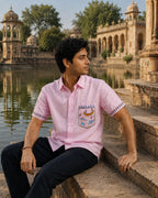 Warli hand-painted pure linen shirt featuring the Matsya pocket artwork of fishermen hunting fish among river creatures, with continuous Tarpa dance painted across both sleeve borders, photographed beside a historic palace reservoir and carved stone architecture.