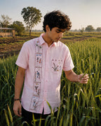 Warli Pravah hand-painted linen shirt featuring a vertical narrative artwork panel composed of palm trees, birds, village figures, basket carriers, florals and shrubs, sun symbols and landscape elements arranged in sequential framed sections beside the button placket. The illustrated pocket artwork depicts flowering plants, a sun motif, ritual figures, a grazing animal and decorative warli borders, observed while inspecting crop yield in a cultivated field during warm natural light.