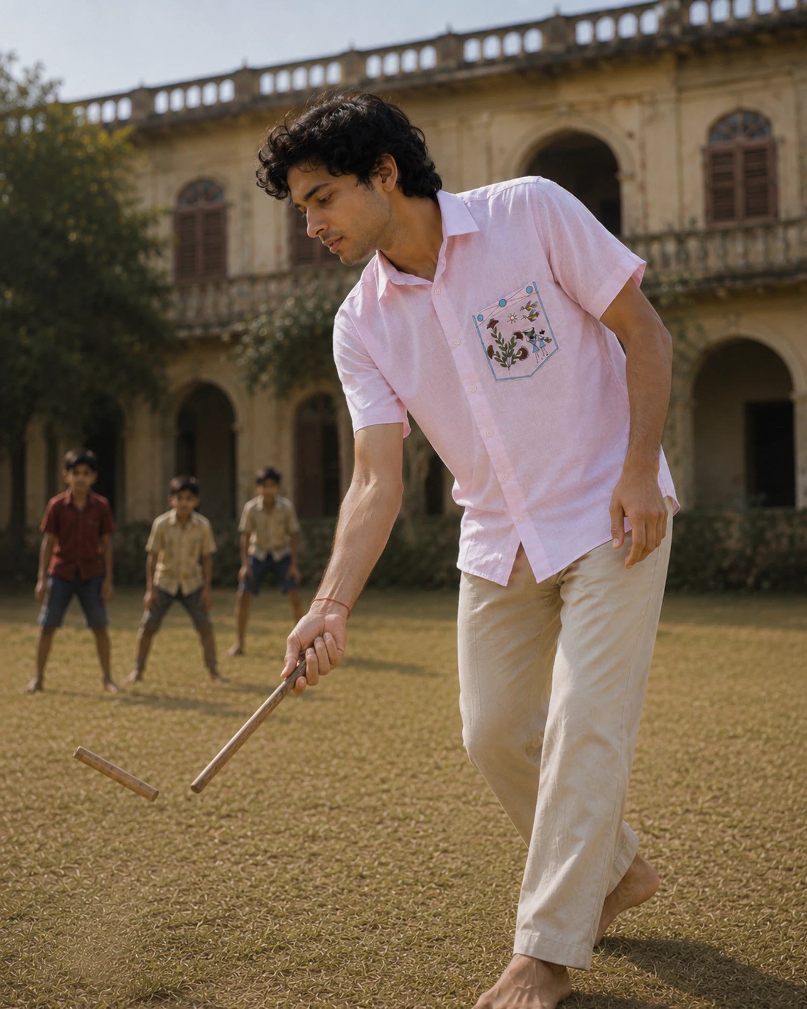 Warli Saanjh hand-painted linen shirt featuring a pocket artwork depicting village life through flowering plants, birds, a sun artwork, and basket-carrying figures, observed during a game of gilli danda in the courtyard of a heritage residence, with village children softly blurred in the background.