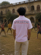 Back view of the Warli Saanjh hand-painted linen shirt featuring a continuous upper-back artwork panel depicting dancers, flowering plants, and village celebrations framed within a traditional warli border, observed in a heritage courtyard as children play gilli danda during a light drizzle.