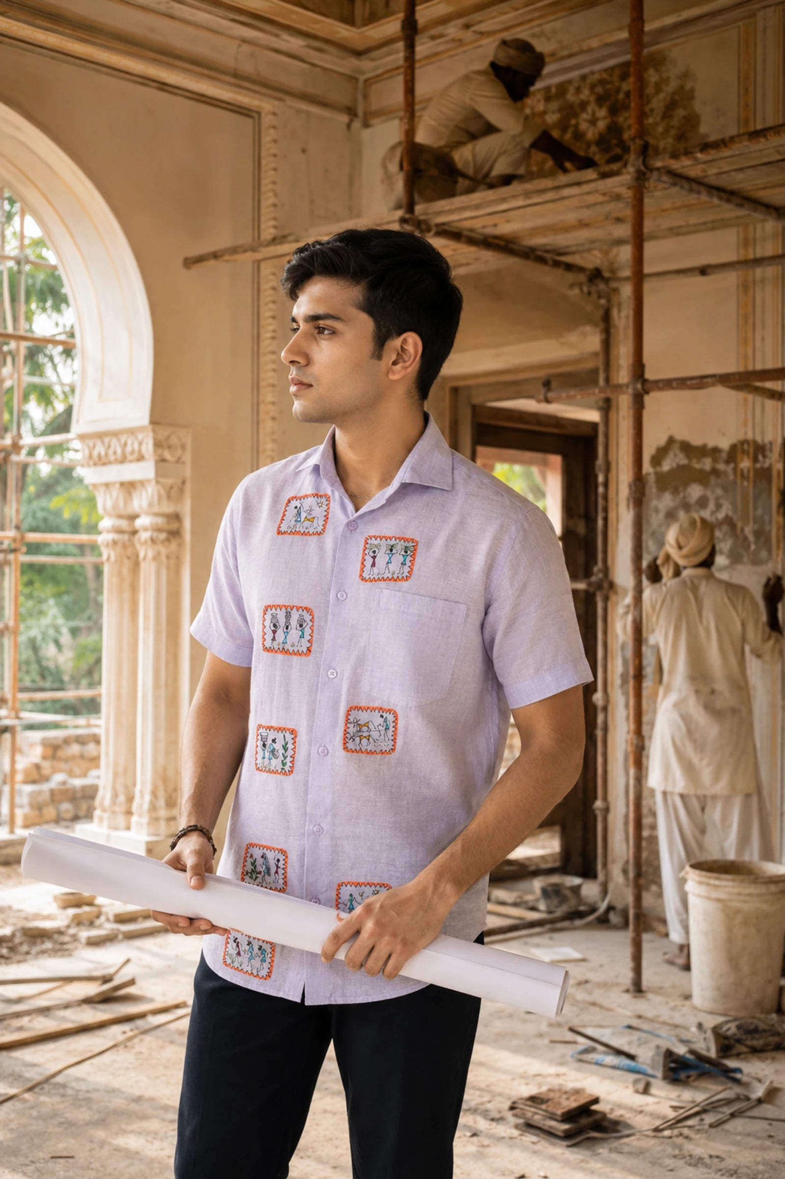 Warli Samriddhi hand-painted linen shirt featuring eight framed Warli narrative panels across the front, worn while observing the restoration of a historic palace interior with artisans and scaffolding in the background, documenting continuity between traditional craftsmanship and architectural preservation.