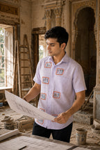Warli Samriddhi hand-painted linen shirt with eight framed Warli narrative artworks across the front, photographed within a heritage palace restoration setting while examining architectural plans.