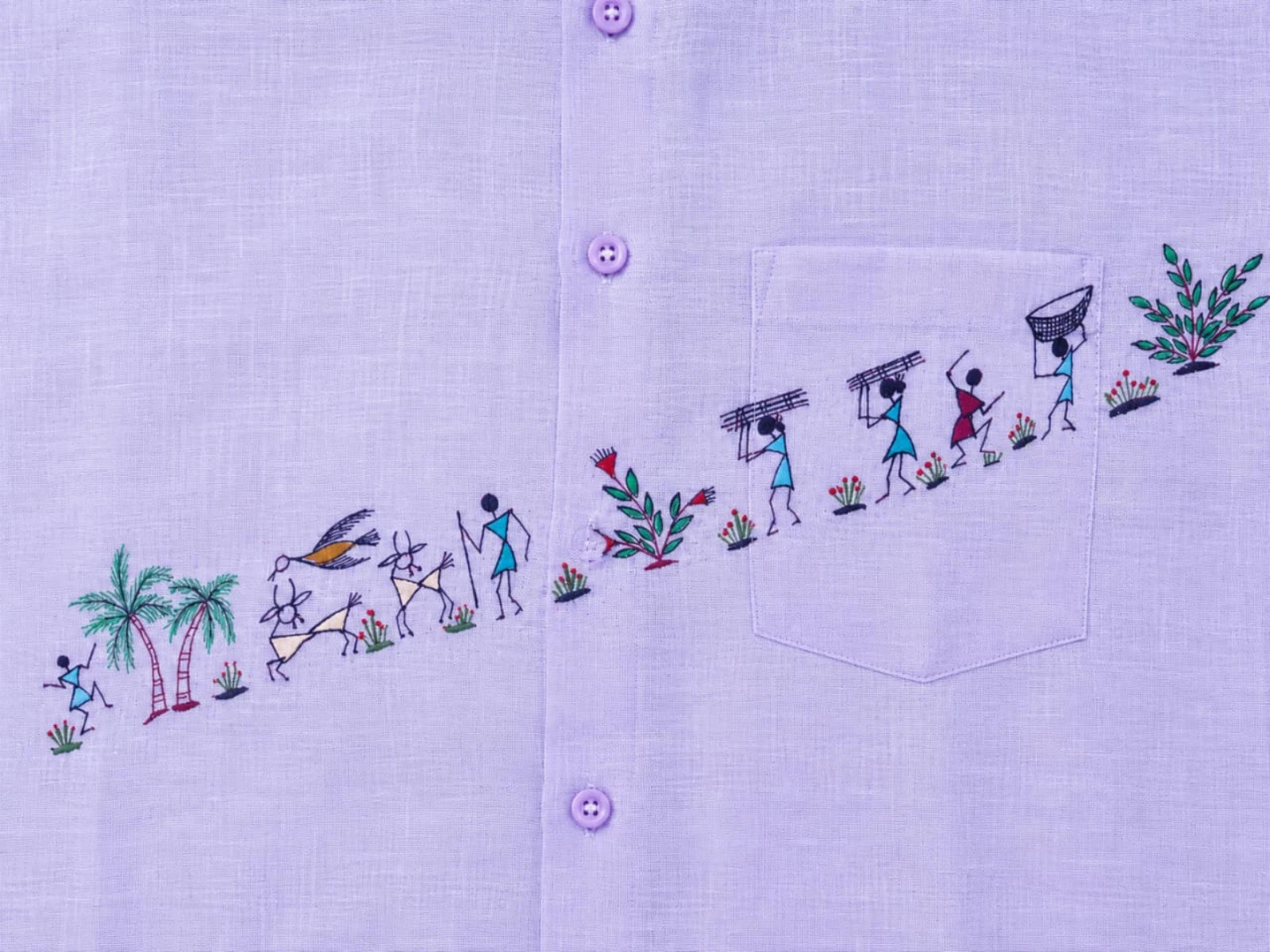Close-up of the diagonal artwork on the Warli Vriddhi hand-painted linen shirt, featuring village figures, palm trees, goats, a bird in flight, flowering foliage, bundle carriers, a joyful dancing figure, a basket carrier, and a leafy shrub arranged in a continuous diagonal composition extending across the front panel, placket, and chest pocket.