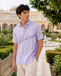 Warli Vriddhi hand-painted linen shirt featuring village figures, palm trees, goats, a bird in flight, flowering foliage, bundle carriers, a joyful dancing figure, a basket carrier, and a leafy shrub rising across the front panel, placket, and chest pocket.