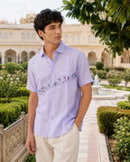 Warli Vriddhi hand-painted linen shirt featuring village figures, palm trees, goats, a bird in flight, flowering foliage, bundle carriers, a joyful dancing figure, a basket carrier, and a leafy shrub rising across the front panel, placket, and chest pocket.