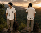 Front and back styled context view of the Padchinh hand-painted linen shirt, featuring a tiger artwork emerging from the lower front panel, botanical detailing above the pocket, and a sequence of paw print artworks along the lower back hem. Photographed on a mountain ridge overlooking a forested valley in warm evening light.