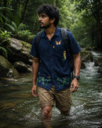 Styled context image of the Vanantara hand-painted pure linen shirt worn while crossing a forest stream, featuring a large butterfly and a smaller butterfly on the upper front chest, a tiger emerging through tall grass beside the chest pocket, and branching bamboo forms with perched birds and butterflies across the lower front panel, shown in a natural woodland setting with flowing water and a field backpack.